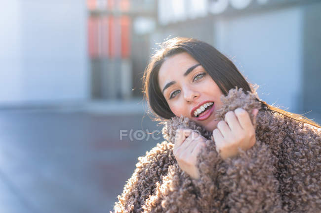 Mujer joven encantadora sonriendo y mirando a la cámara mientras se envuelve en abrigo cálido y elegante y de pie sobre el fondo borroso de la calle de la ciudad en el día soleado - foto de stock