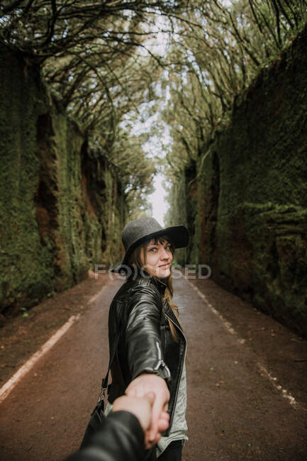 Вид сбоку на элегантную даму в шляпе и кожаной куртке, держащую человека за руку и стоящую на тропинке между мрачной аллеей высоких стен и лесов — стоковое фото