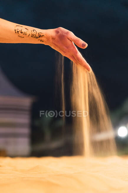 Татуйована рука анонімної людини, що проливає сухий пісок на розмитому тлі міста — стокове фото