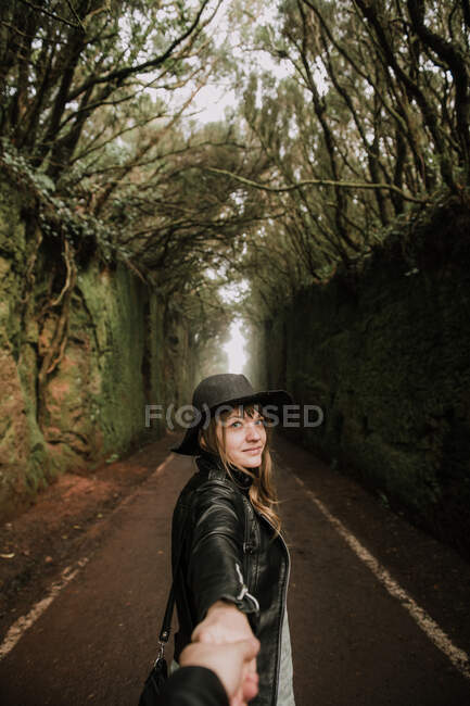 Vista laterale di elegante signora in cappello e giacca di pelle che tiene la mano della persona e in piedi sul sentiero tra vicolo buio di alte pareti e boschi — Foto stock