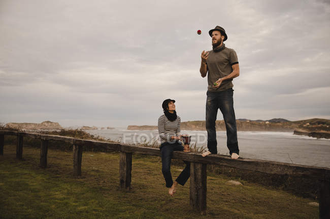 Young guy in hat juggling balls near elegant woman in cap with ethic drum sitting on seat near coast of sea and cloudy sky — Stock Photo