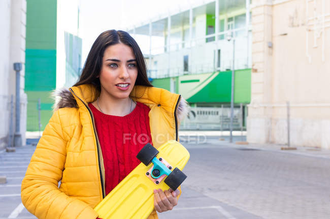 Bella giovane donna in abito alla moda in possesso di skateboard giallo e guardando altrove mentre in piedi sulla strada della città nella giornata di sole — Foto stock
