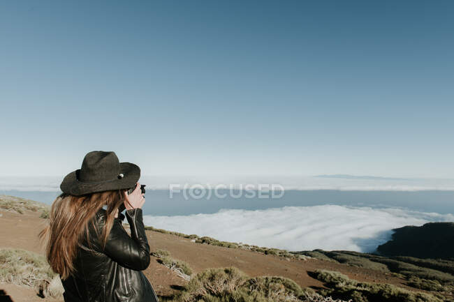 Vista posterior de la mujer con cámara de fotos de pie en la cima de la colina y mirando el paisaje nublado - foto de stock