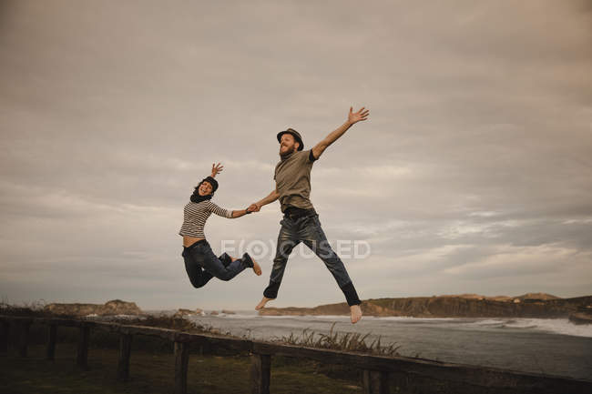 Young happy woman and man in hats holding hands and having fun on seat on coast near waving sea and cloudy sky — Stock Photo
