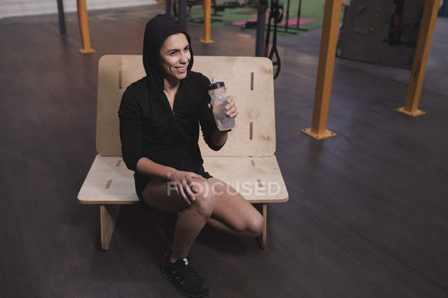 Woman in sportswear with bottle of water sitting on bench in gym — Stock Photo