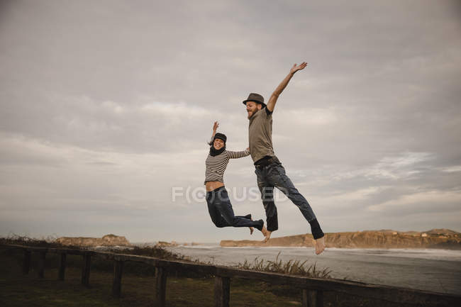 Молодые счастливые женщина и мужчина в шляпах, держась за руки и веселясь на берегу возле машущего морем и облачным небом — стоковое фото