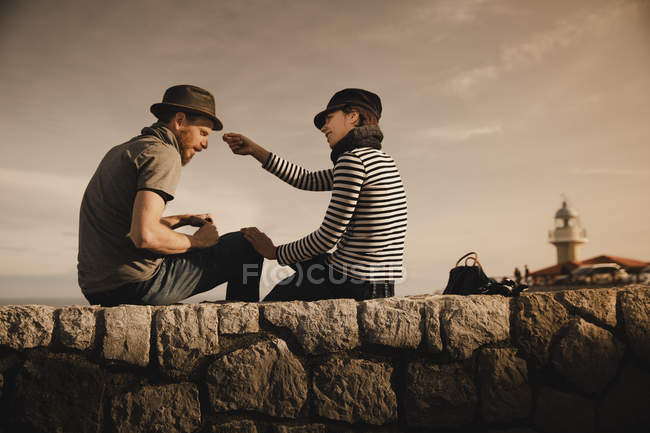 Вид сбоку стильной элегантной пары, сидящей на скалах рядом с маяком и прекрасным небом — стоковое фото
