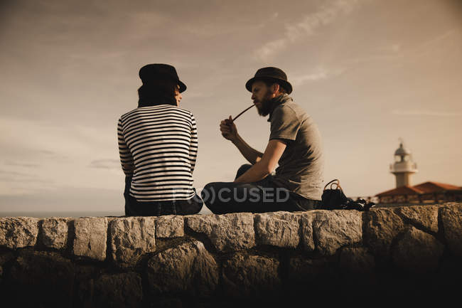 Vista lateral do cara elegante em cachimbo de fumar chapéu e mulher elegante em volume de leitura de boné e sentado em rochas perto de farol — Fotografia de Stock
