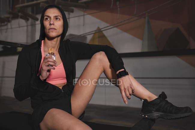 Jeune femme en vêtements de sport avec bar énergétique assis sur le banc dans la salle de gym — Photo de stock