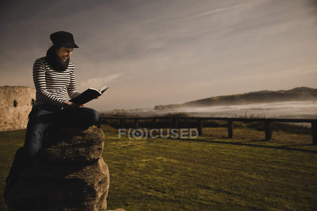 Elegante mujer con gorra sentada en el asiento en la costa cerca del mar ondulante - foto de stock