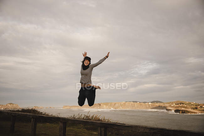 Jovem mulher de chapéu se divertindo no assento na costa perto do mar ondulando e céu nublado — Fotografia de Stock