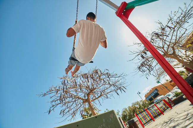Vista trasera del hombre descalzo en columpios divertirse y relajarse en el parque infantil de la ciudad a la luz del sol - foto de stock