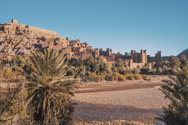 Каменные сооружения в старом городе возле зеленых деревьев на берегу реки и голубого неба в Марракеше, Марокко — стоковое фото