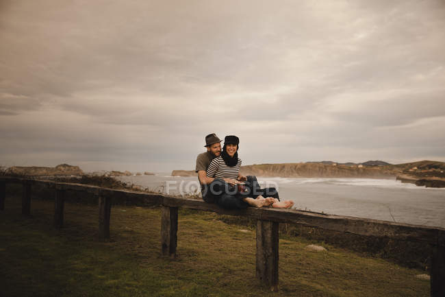 Jeune homme en chapeau près de femme élégante jouant sur le tambour main en chapeau sur le siège près de la côte de la mer et ciel nuageux — Photo de stock