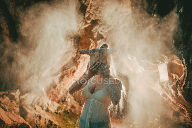 Woman wearing white lace top covering face with animal skull while standing in flying dry dust inside of cave — Stock Photo