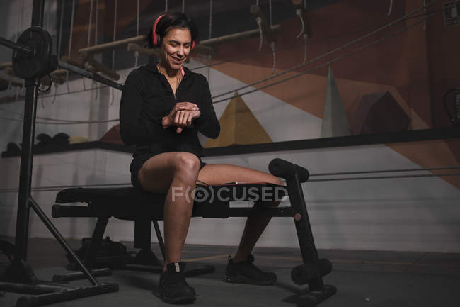 Frau in Sportbekleidung kontrolliert Uhrzeit und sitzt auf Bank im Fitnessstudio — Stockfoto