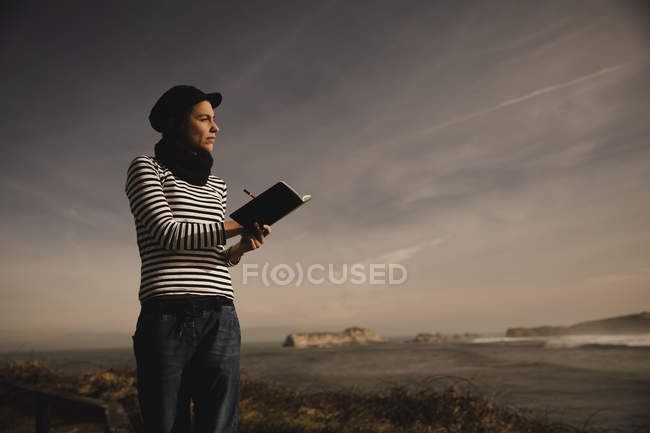Элегантная женщина в кепке делает заметки в блокноте на сиденье на берегу возле машущего морем — стоковое фото