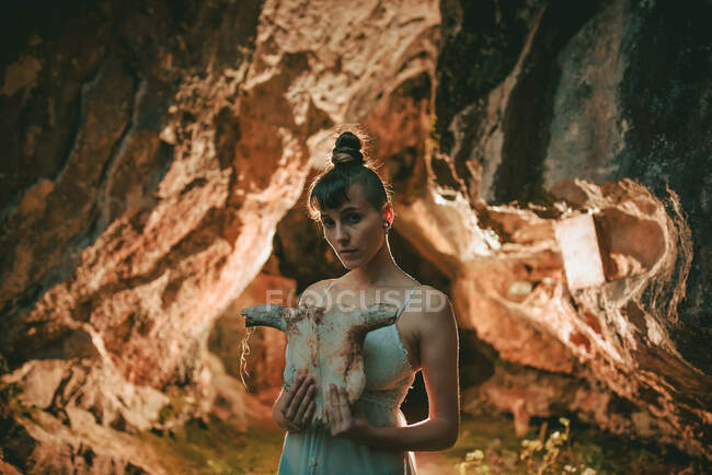 Mulher vestindo top de renda branca segurando crânio animal enquanto estava em pé em pó seco voando dentro da caverna — Fotografia de Stock