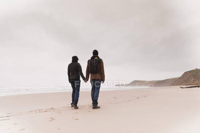 Rückenansicht eines jungen Paares mit Rucksack in warmer Kleidung, das sich am Sandstrand in der Nähe von Meer und Hügeln die Hände hält — Stockfoto