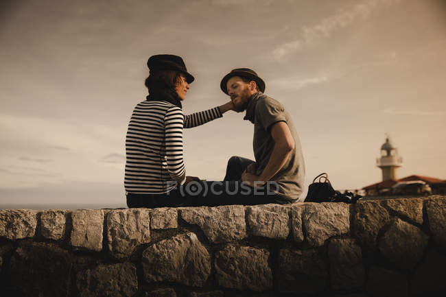 Vista lateral do elegante casal elegante sentado em rochas perto de farol e céu maravilhoso — Fotografia de Stock