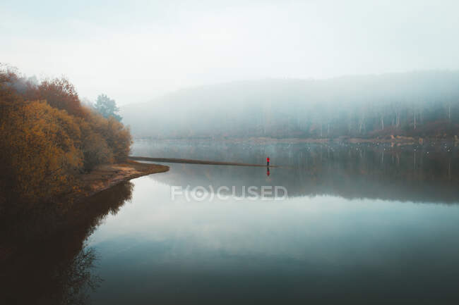 Pintoresco paisaje de hermoso lago silencioso con reflejo de bosque y turista solitario en espesa niebla - foto de stock