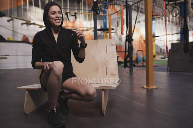 Woman in sportswear with energy bar sitting on bench in gym — Stock Photo
