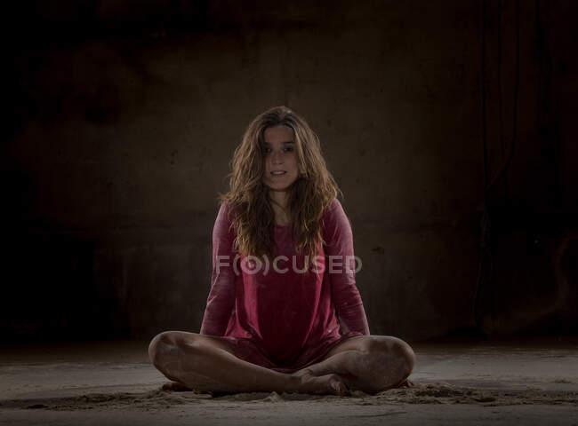 Молодая уставшая леди в красном платье сидит в позе лотоса между песком и смотрит в камеру в темной комнате — стоковое фото
