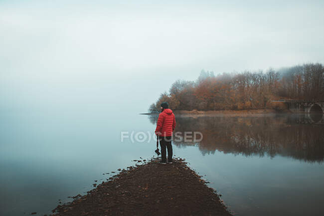 Visão traseira do fotógrafo em casaco vermelho em pé na costa do lago tranquilo na névoa — Fotografia de Stock