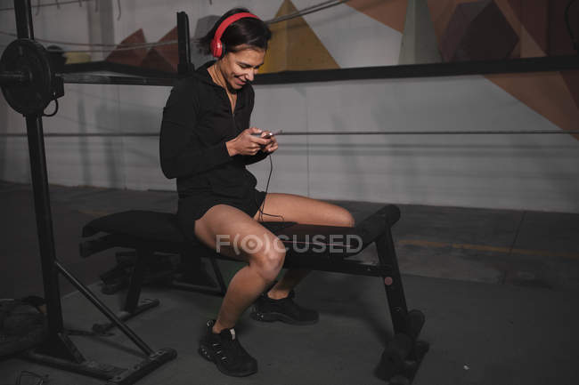 Side view of woman in sportswear with headphones and gadget listening music and sitting in gym — Stock Photo