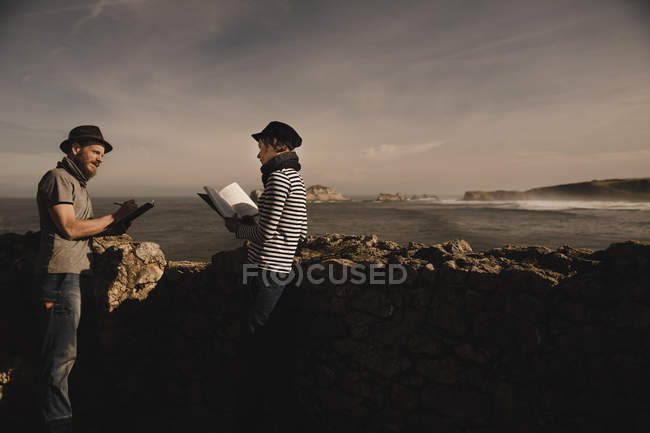 Cara elegante em chapéu e mulher elegante em volume de leitura de boné perto de rochas na costa do mar e céu maravilhoso — Fotografia de Stock