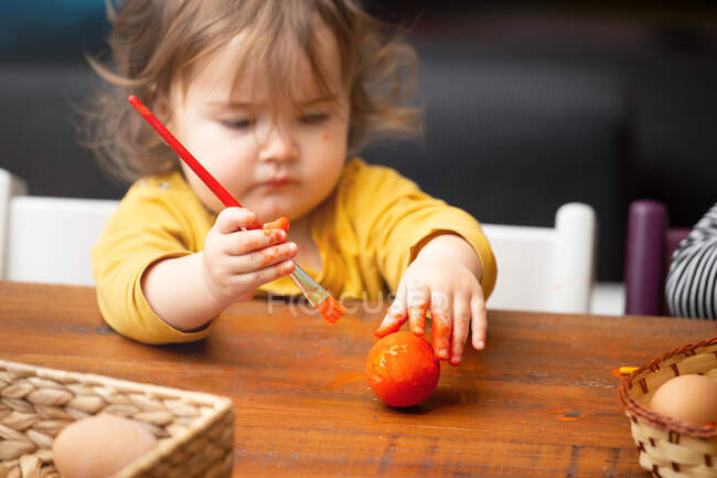 Очаровательная девочка использует кисть, чтобы нарисовать пасхальное яйцо, сидя за столом. — стоковое фото