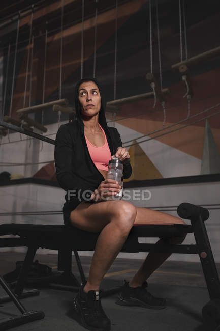 Mujer en ropa deportiva con botella de agua sentada en el banco en el gimnasio - foto de stock