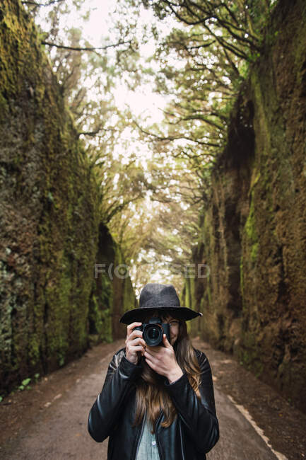 Элегантная леди в шляпе и кожаной куртке, снимающая на камеру и стоящая на тропинке между темными аллеями высоких стен и лесов — стоковое фото