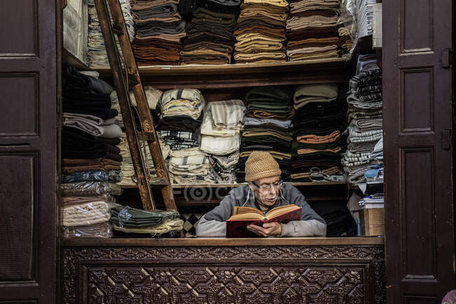31 décembre 2017 - Marrakech, Maroc : Vendeur adulte assis au comptoir avec tissu et livre de lecture — Photo de stock