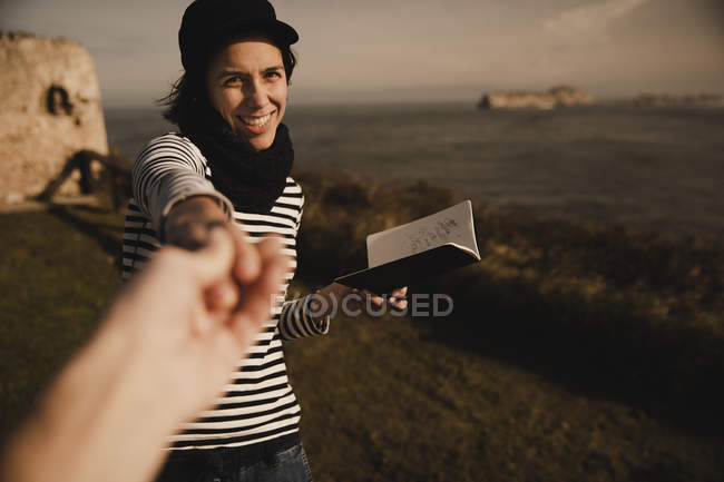 Élégante femme gaie en bonnet avec bloc-notes tenant la main de l'homme sur la côte près de la mer — Photo de stock