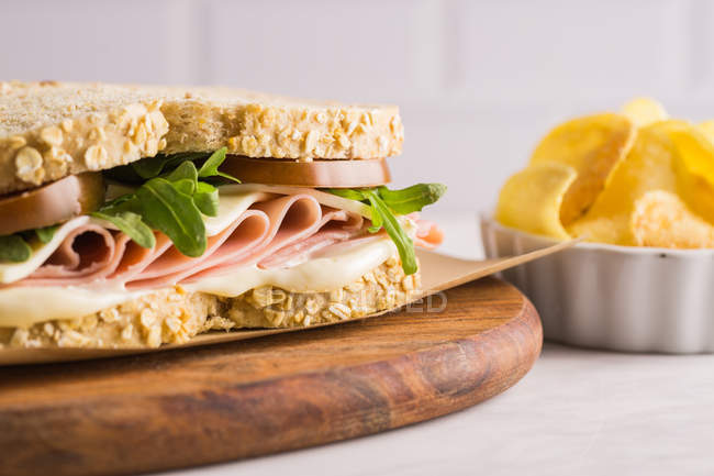 Delicious sandwich with ham, cheese and greens on wooden chopping board with potato chips on white background — Stock Photo