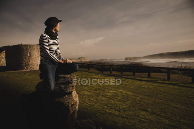 Femme élégante en casquette assise sur le siège sur la côte près de la mer ondulante — Photo de stock
