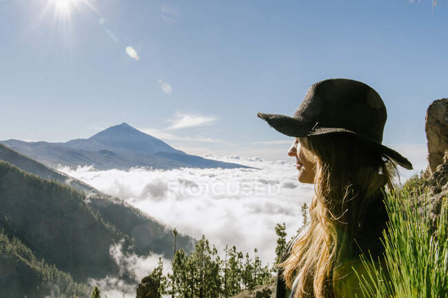 Vista lateral de la mujer con sombrero de pie en la cima de la colina mirando el bosque y el paisaje nublado en un día soleado - foto de stock