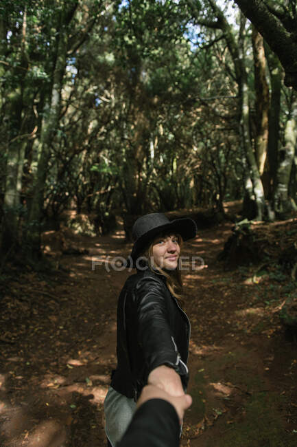 З боку погляд на елегантну леді в капелюсі і шкіряний піджак, тримаючи за руку людину і стоячи на стежці між муром алеї високих стін і лісу. — стокове фото