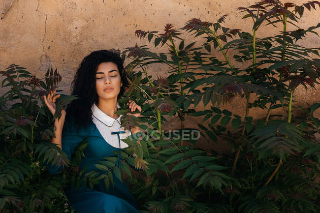 Atractiva mujer árabe en vestido entre plantas cerca de la pared - foto de stock