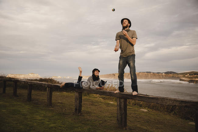 Молодой парень в шляпе жонглирует шарами возле элегантной женщины в шапочке с этичным барабаном, сидящим на сиденье у берегов моря и облачного неба — стоковое фото