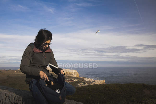 Donna elegante tirando fuori un libro dallo zaino e seduto sul prato vicino faro sulla riva e cielo meraviglioso — Foto stock