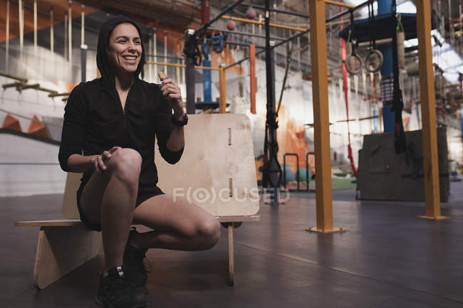Young woman in sportswear with energy bar sitting on bench in gym — Stock Photo