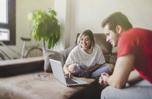 Souriant jeune homme et femme assis sur le canapé et ordinateur portable de navigation dans le salon — Photo de stock