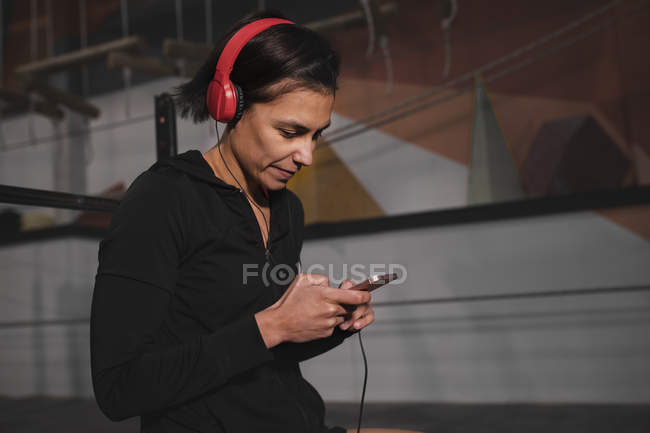 Вид збоку на жінку в спортивному одязі з навушниками та гаджетом, що слухає музику та сидить у спортзалі — стокове фото