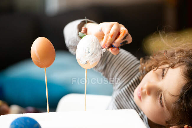 Doce menina focada usando escova para pintar frágeis ovos de Páscoa em paus no fundo turvo do quarto em casa — Fotografia de Stock