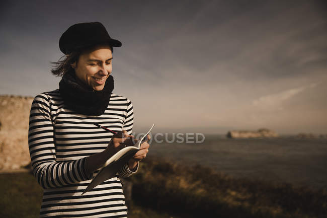 Elegant woman in cap taking notes in notepad on seat on coast near waving sea — Stock Photo