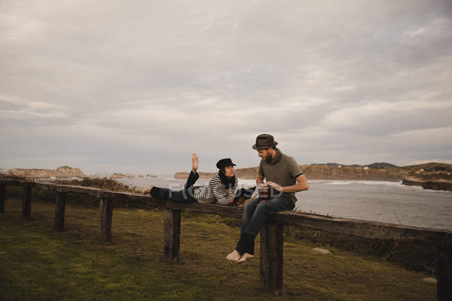 Jovem de chapéu tocando na mão tambor perto de mulher elegante em boné com bloco de notas e lápis deitado no assento perto da costa do mar — Fotografia de Stock