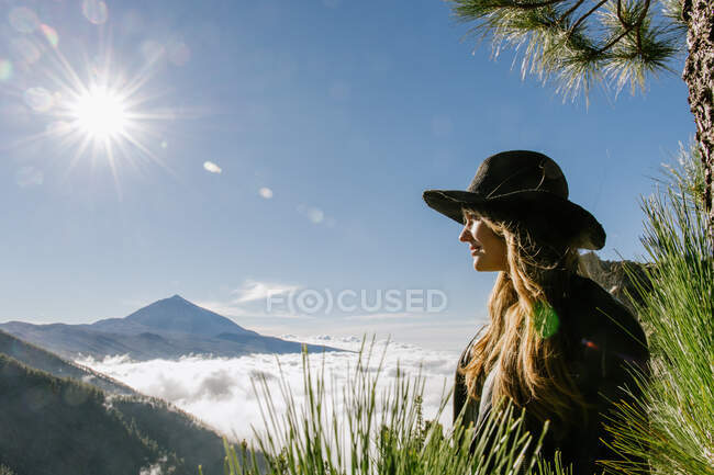 Vista lateral de la mujer con sombrero de pie en la cima de la colina mirando el bosque y el paisaje nublado en un día soleado - foto de stock