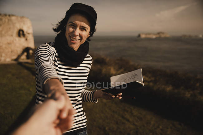 Elegant cheerful woman in cap with notepad holding crop hand of person on coast near sea — Stock Photo